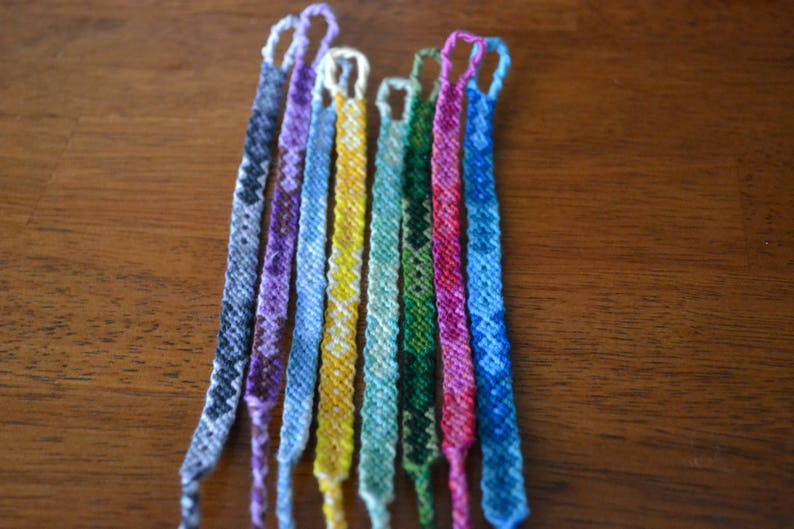 Pode incluir: Uma cole&ccedil;&atilde;o de dez pulseiras de amizade coloridas, cada uma com um padr&atilde;o &uacute;nico. As pulseiras s&atilde;o feitas com diferentes tons de azul, roxo, verde, amarelo e vermelho.