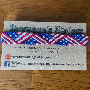Puede incluir: Pulsera de la amistad roja, blanca y azul con un patrón diagonal. La pulsera está en una tarjeta que dice "Suzanne's Strings" y "¡La amistad hace girar el mundo!"