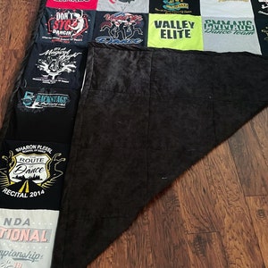 May include: A patchwork quilt constructed from assorted t-shirts, with a black reverse side. The t-shirts showcase text and graphics associated with dance, including team names such as "Emmaus Dance Team" and "Valley Elite". The quilt is displayed on a wooden floor.