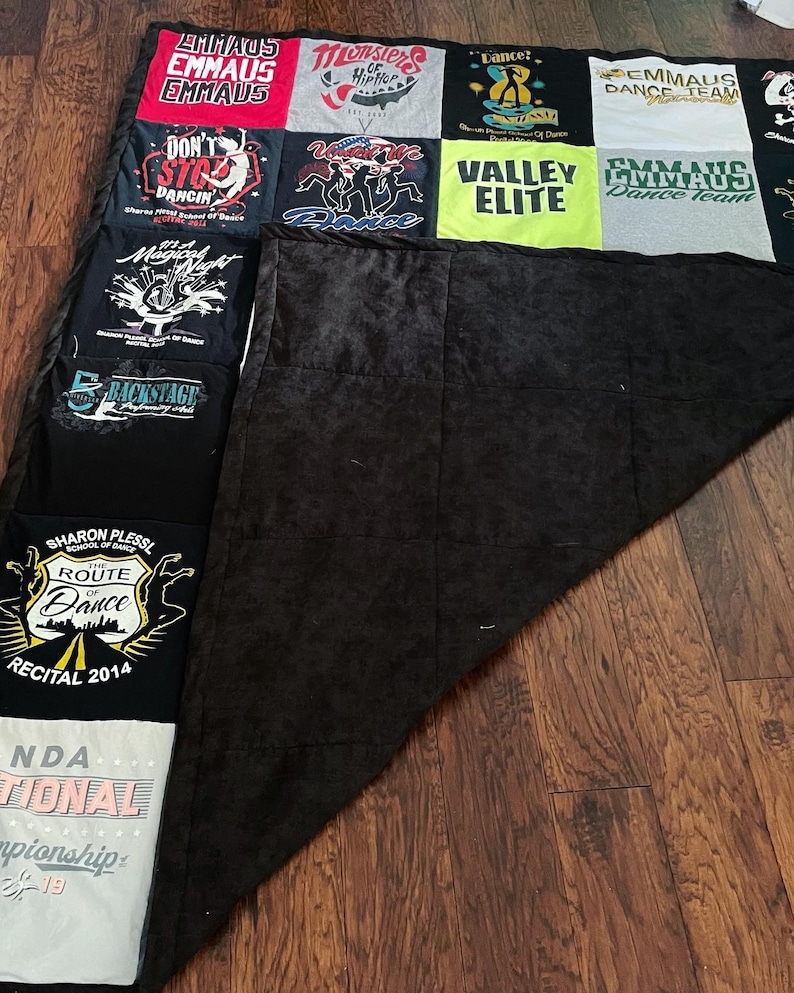 May include: A patchwork quilt constructed from assorted t-shirts, with a black reverse side. The t-shirts showcase text and graphics associated with dance, including team names such as "Emmaus Dance Team" and "Valley Elite". The quilt is displayed on a wooden floor.