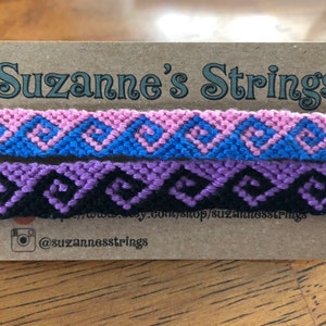 May include: Two friendship bracelets with geometric patterns. The top bracelet is pink, blue, and white. The bottom bracelet is purple, black, and white. The bracelets are displayed on a brown card with the text "Suzanne's Strings" and a social media handle.