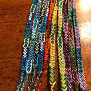 Pode incluir: Uma cole&ccedil;&atilde;o de pulseiras de amizade coloridas com v&aacute;rios padr&otilde;es geom&eacute;tricos. As pulseiras s&atilde;o feitas com fio de bordar colorido e s&atilde;o amarradas juntas nas pontas.