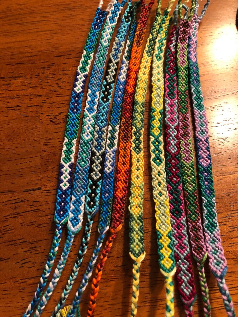 Pode incluir: Uma cole&ccedil;&atilde;o de pulseiras de amizade coloridas com v&aacute;rios padr&otilde;es geom&eacute;tricos. As pulseiras s&atilde;o feitas com fio de bordar colorido e s&atilde;o amarradas juntas nas pontas.