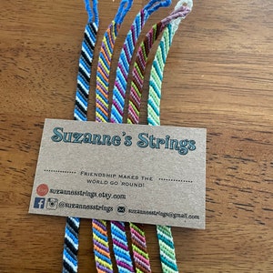 Può includere: Un set di sei braccialetti dell'amicizia colorati con un motivo a chevron. I braccialetti sono esposti su una carta marrone con il testo "Suzanne's Strings" e lo slogan "L'amicizia fa girare il mondo!"