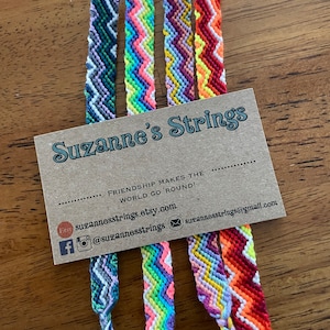 May include: A collection of colorful friendship bracelets with a chevron pattern. The bracelets are made with embroidery floss and are tied with a knot at the end. The bracelets are displayed on a brown card with the text "Suzanne's Strings" and the phrase "Friendship makes the world go 'round!"