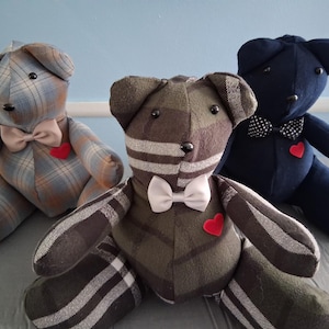 May include: Three stuffed teddy bears in a seated position. The left bear has a blue and brown plaid pattern, the center bear has a dark green and black plaid pattern, and the right bear is solid navy blue. Each bear features a bow tie and a red heart.