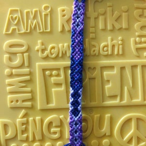 Pode incluir: Um bracelete de amizade feito com fio azul, roxo e branco. O bracelete est&aacute; sobre uma superf&iacute;cie amarela com a palavra "amigo" em relevo.
