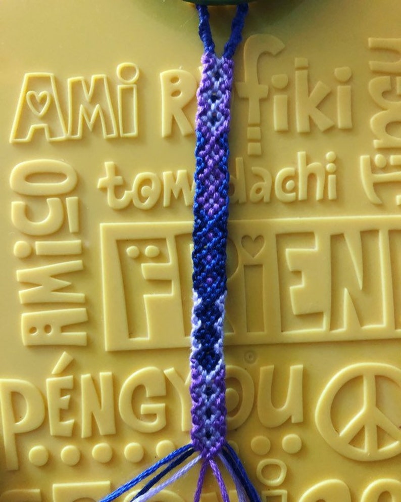 Pode incluir: Um bracelete de amizade feito com fio azul, roxo e branco. O bracelete est&aacute; sobre uma superf&iacute;cie amarela com a palavra "amigo" em relevo.
