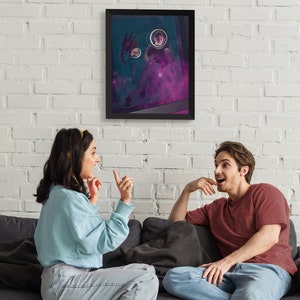 May include: Two people are sitting on a couch and looking at a framed print on the wall. The print is a dark blue background with a person in a purple haze and three circular images of people. The people on the couch are wearing casual clothing.