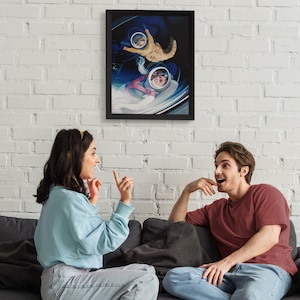 May include: A framed art print depicting a cat and a person in astronaut helmets floating in a spaceship. The artwork is in a black frame and hangs on a white brick wall. The image is a surreal, space-themed illustration.