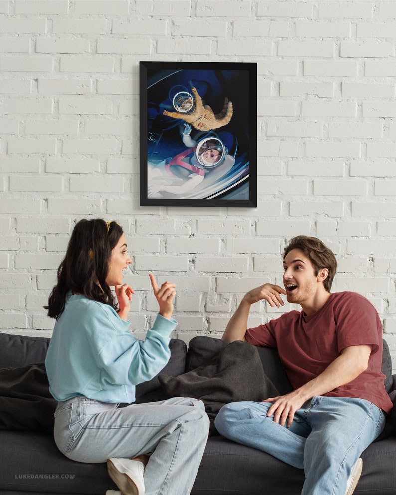 May include: A framed art print depicting a cat and a person in astronaut helmets floating in a spaceship. The artwork is in a black frame and hangs on a white brick wall. The image is a surreal, space-themed illustration.