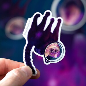 May include: A sticker of a cat wearing an astronaut helmet, floating in space. The cat is upside down, with its tail pointing towards the bottom of the sticker. The background is a purple and blue nebula.