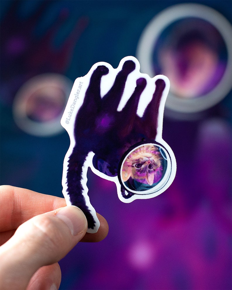 May include: A sticker of a cat wearing an astronaut helmet, floating in space. The cat is upside down, with its tail pointing towards the bottom of the sticker. The background is a purple and blue nebula.