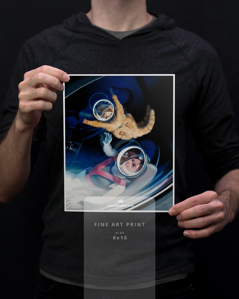May include: A fine art print featuring an astronaut and a cat in space. The artwork is a vibrant illustration with a blue and white color scheme. The print is 8x10 inches in size and includes the text "FINE ART PRINT" and the artist's website.
