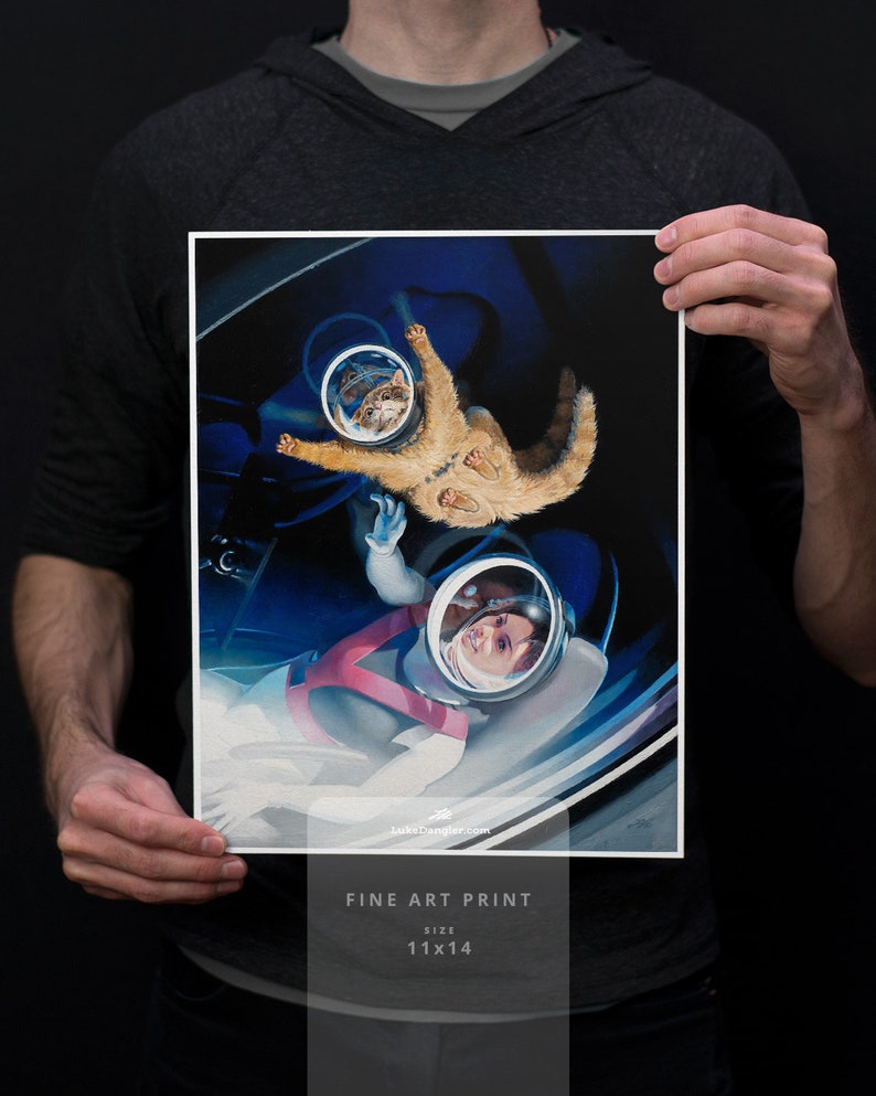 May include: Fine art print featuring a whimsical space scene with a cat and a person in astronaut helmets. The print is 11x14 inches and includes the text "Fine Art Print" and "Size 11x14". The artwork has a blue and white color scheme.