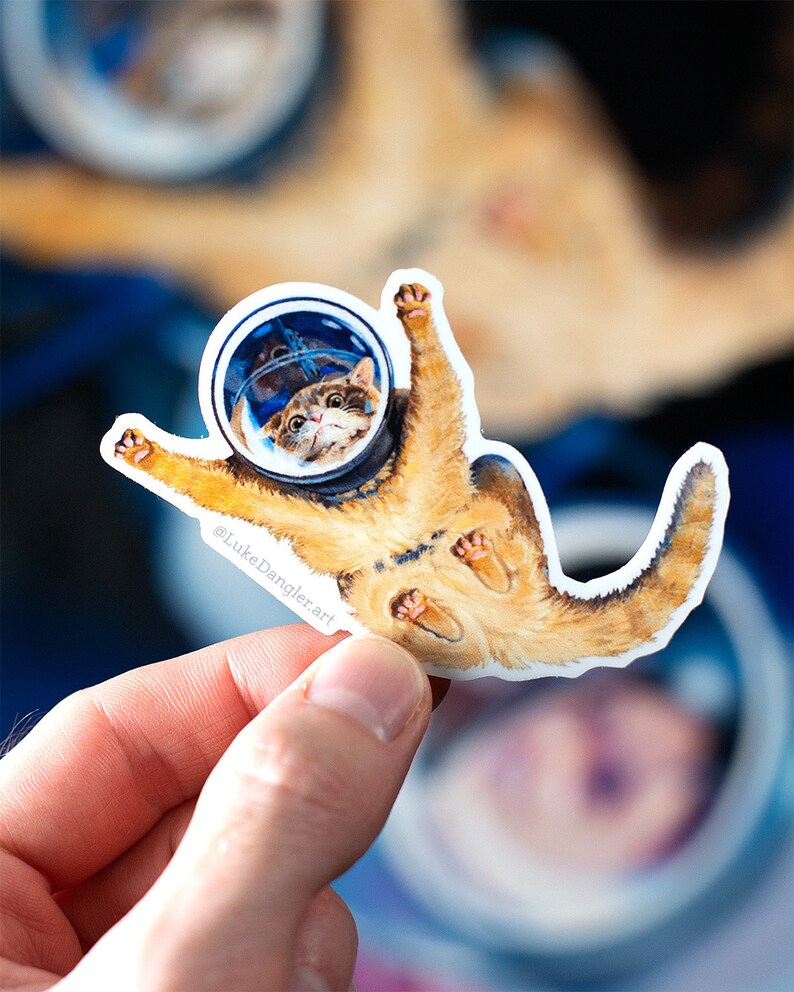 May include: A sticker featuring a whimsical illustration of a cat in an astronaut helmet, floating in space. The cat is orange and white, with its paws outstretched. The sticker has a white border and the artist's signature, "@LukeDanglerart."