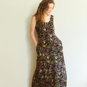May include: A sleeveless, floor-length black dress with a vibrant floral pattern. The dress features a scoop neckline and pockets. The colorful floral print includes red, yellow, and green accents. The dress is made of a lightweight fabric.