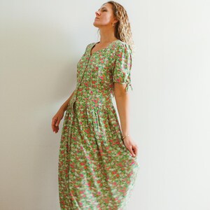 Op de afbeelding: Een midi-jurk met een dicht patroon van kleine rode, witte en roze bloemen op een felgroene ondergrond. Het kledingstuk heeft korte mouwen met strikdetails, een V-hals, een knoopsluiting aan de voorzijde en een geplooide taille met een wijde rok.