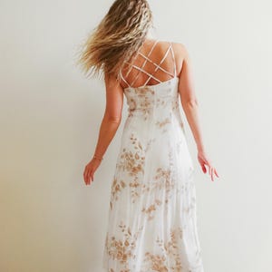 Puede incluir: Vestido blanco con estampado floral en tonos marrones. El vestido presenta un diseño de espalda cruzada y tirantes finos. La modelo lleva el vestido y tiene el pelo largo y rubio.