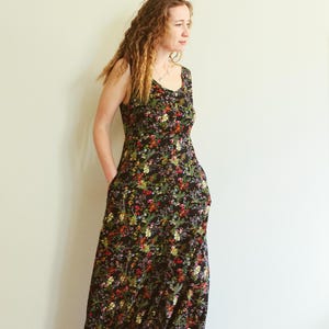 May include: A sleeveless, floor-length dress with a black background and a vibrant floral pattern in red, yellow, and green. The dress has a scoop neckline and pockets. The model is wearing the dress.