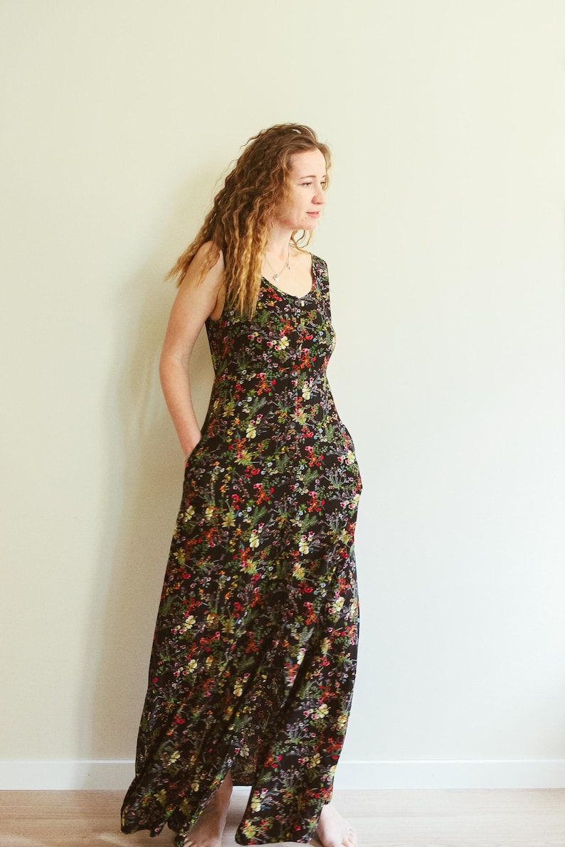 May include: A sleeveless, floor-length dress with a black background and a vibrant floral pattern in red, yellow, and green. The dress has a scoop neckline and pockets. The model is wearing the dress.