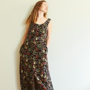 May include: A sleeveless black floral maxi dress with a slit. The dress features a scoop neckline and pockets. The floral pattern includes red, yellow, and green flowers. The dress is worn by a person.
