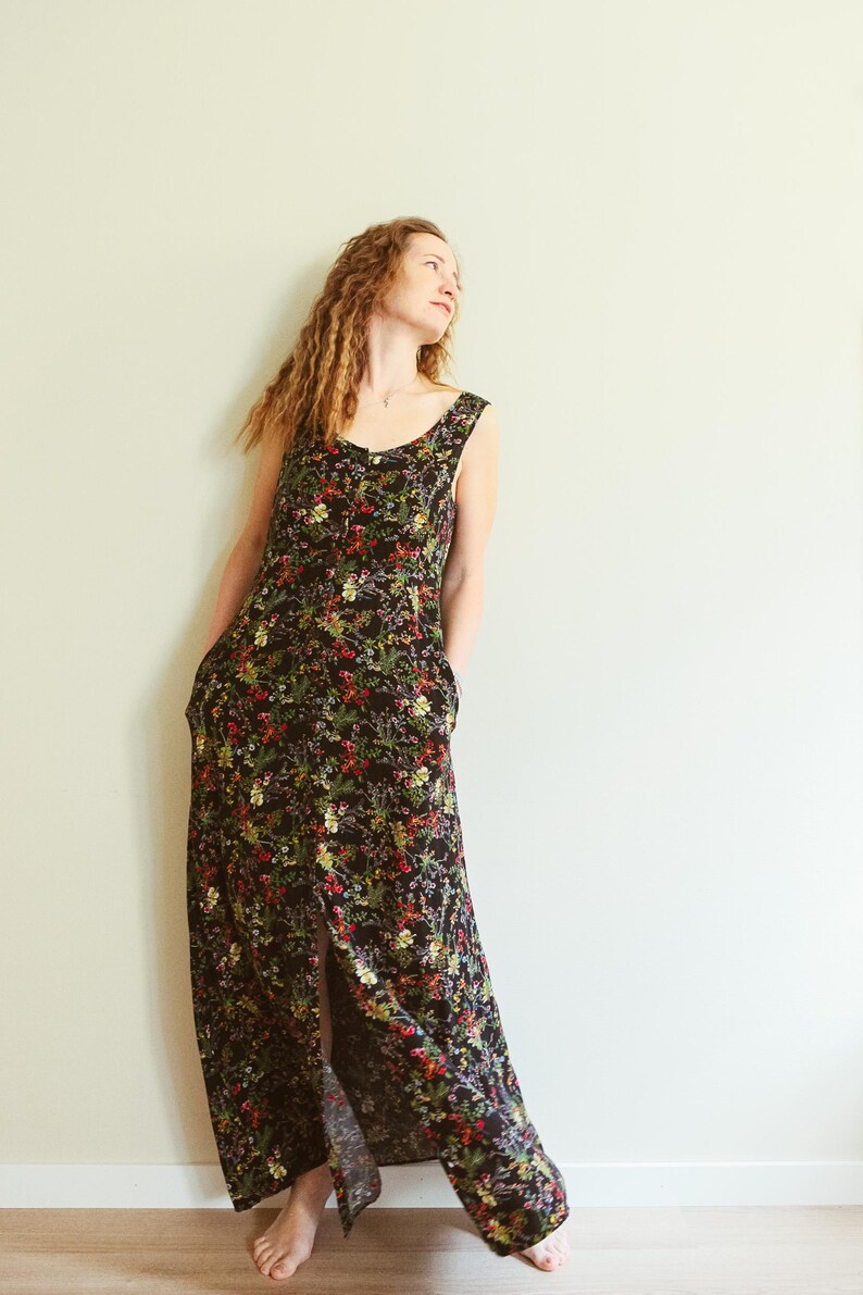 May include: A sleeveless black floral maxi dress with a slit. The dress features a scoop neckline and pockets. The floral pattern includes red, yellow, and green flowers. The dress is worn by a person.