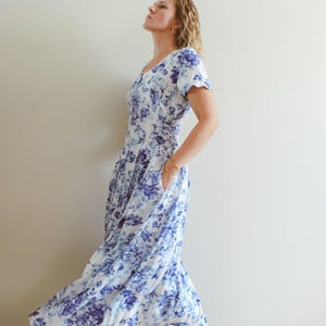 Könnte beinhalten: Ein wadenlanges Kleid mit blauem Blumenmuster auf weißem Grund. Das Kleidungsstück hat kurze Ärmel, einen V-Ausschnitt, ein körpernahes Oberteil und einen ausgestellten Rock mit Seitentaschen. Der Stoff zeigt eine fließende Silhouette.
