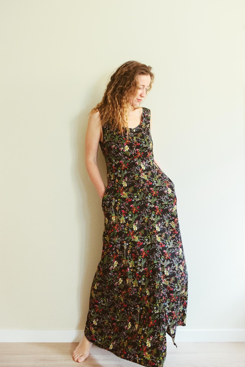 May include: A sleeveless, floor-length black dress with a vibrant floral pattern. The dress features a scoop neckline and pockets. The colorful floral print includes red, yellow, and green accents. The dress is made of a lightweight fabric.