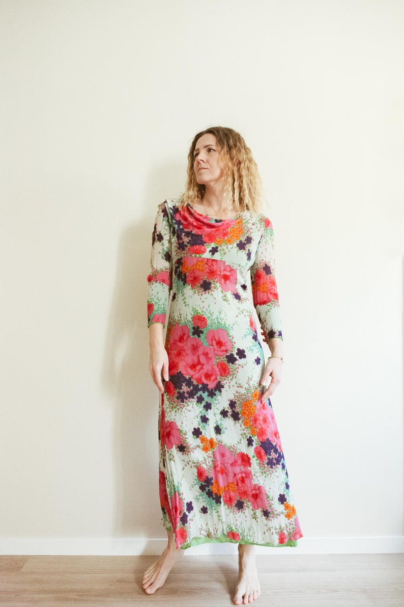 May include: A long, flowing maxi dress with a floral print in shades of pink, orange, green, and purple. The dress has a cowl neck and three-quarter sleeves.