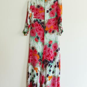 May include: A long, flowing maxi dress with a floral print. The dress has a green background with pink, orange, and purple flowers. The dress has a V-neck and long sleeves.