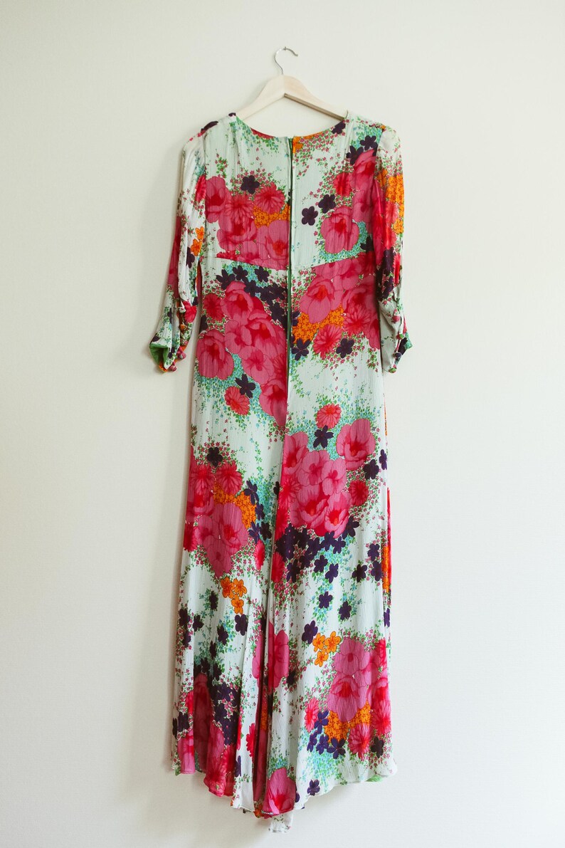 May include: A long, flowing maxi dress with a floral print. The dress has a green background with pink, orange, and purple flowers. The dress has a V-neck and long sleeves.