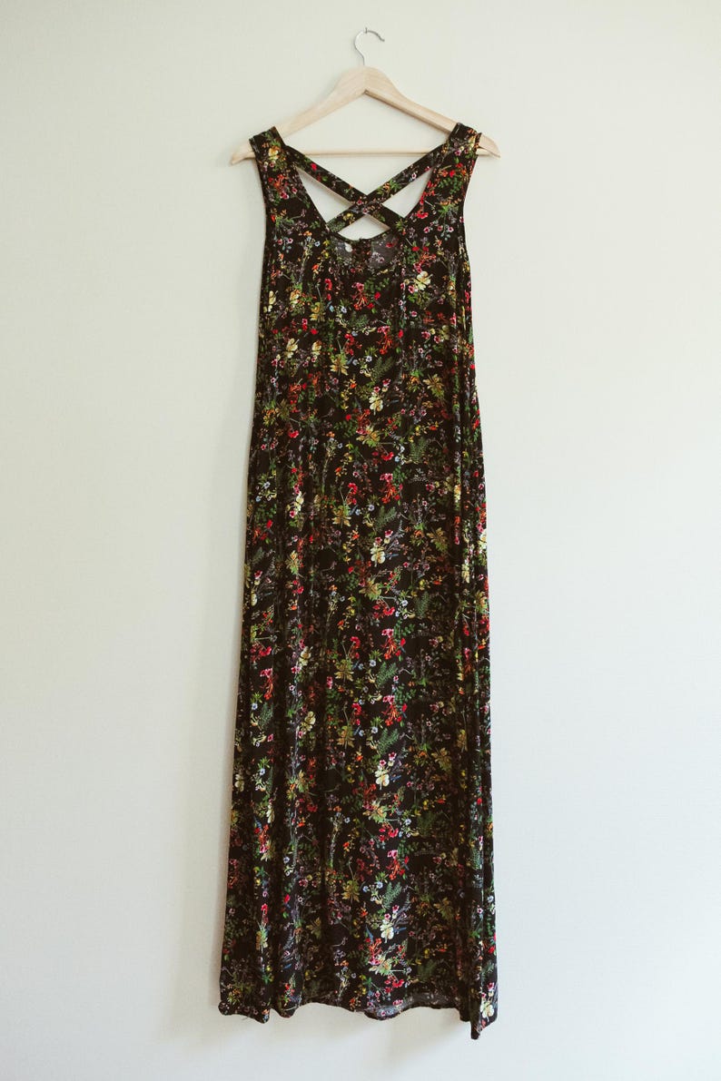 May include: A black maxi dress with a floral pattern in red, yellow, and green. The dress has a crisscross back design and hangs on a wooden hanger against a white wall.