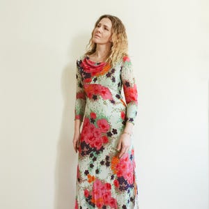 May include: A woman wearing a long, flowing floral dress with a cowl neck. The dress is a mix of pink, red, orange, green, and white flowers on a light green background.