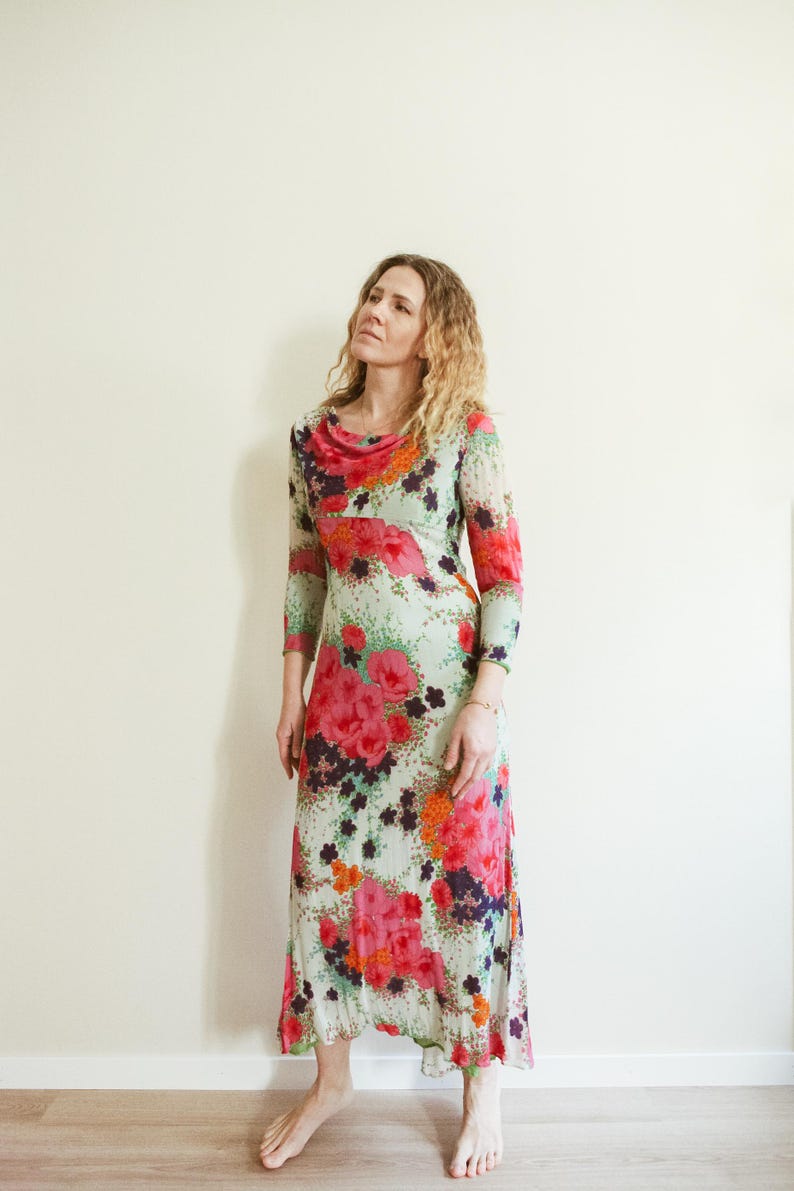 May include: A woman wearing a long, flowing floral dress with a cowl neck. The dress is a mix of pink, red, orange, green, and white flowers on a light green background.