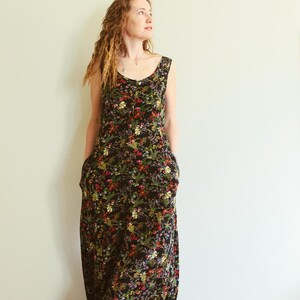 May include: A sleeveless black maxi dress with a vibrant floral pattern in red, yellow, and green. The dress features a scoop neckline and pockets. The model is standing in front of a plain white wall.