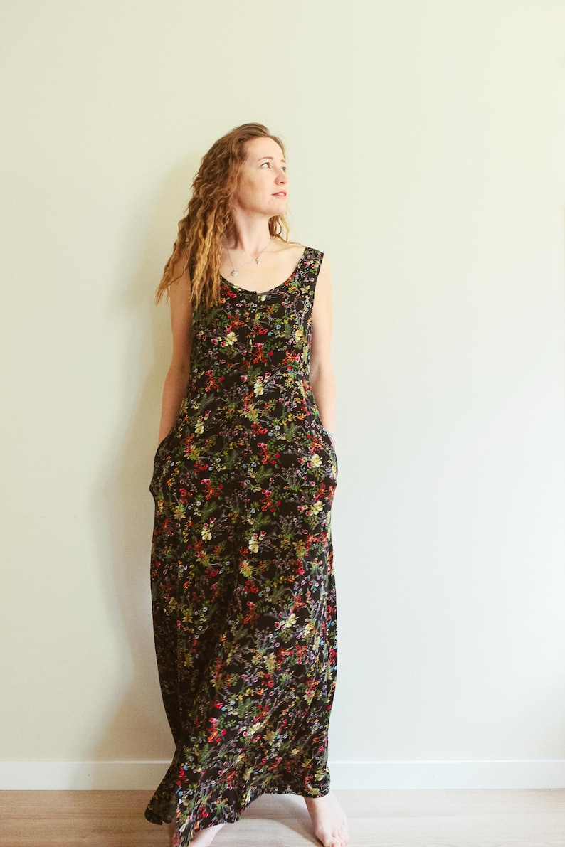 May include: A sleeveless black maxi dress with a vibrant floral pattern in red, yellow, and green. The dress features a scoop neckline and pockets. The model is standing in front of a plain white wall.