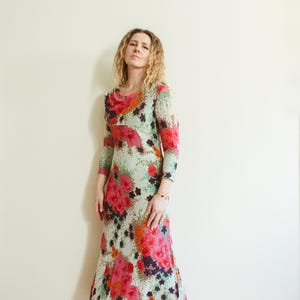 May include: A woman wearing a long, flowing floral dress with a green, pink, and white background. The dress has a scoop neck and long sleeves. The woman is standing in front of a white wall.