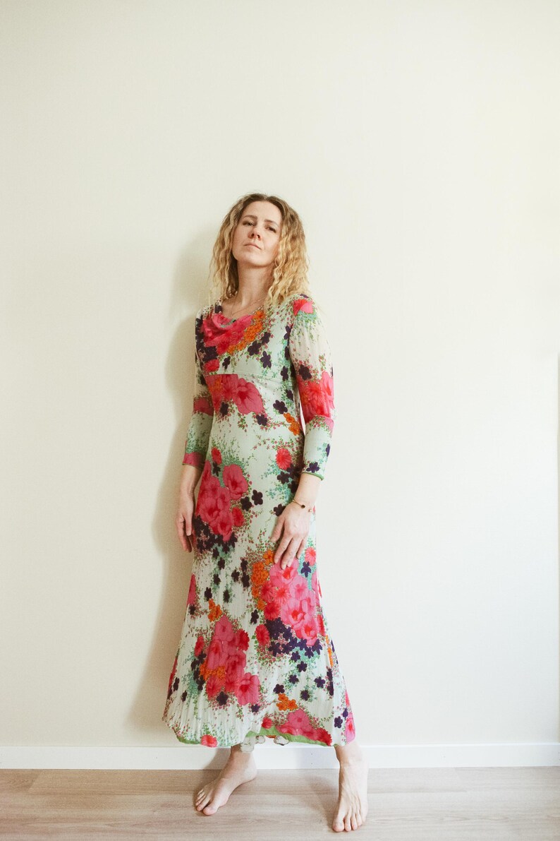 May include: A woman wearing a long, flowing floral dress with a green, pink, and white background. The dress has a scoop neck and long sleeves. The woman is standing in front of a white wall.