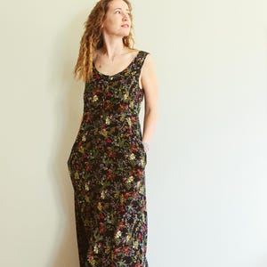 May include: A sleeveless, floor-length dress with a black background and a vibrant floral pattern in red, yellow, and green. The dress has a scoop neckline and pockets. The model is wearing the dress.
