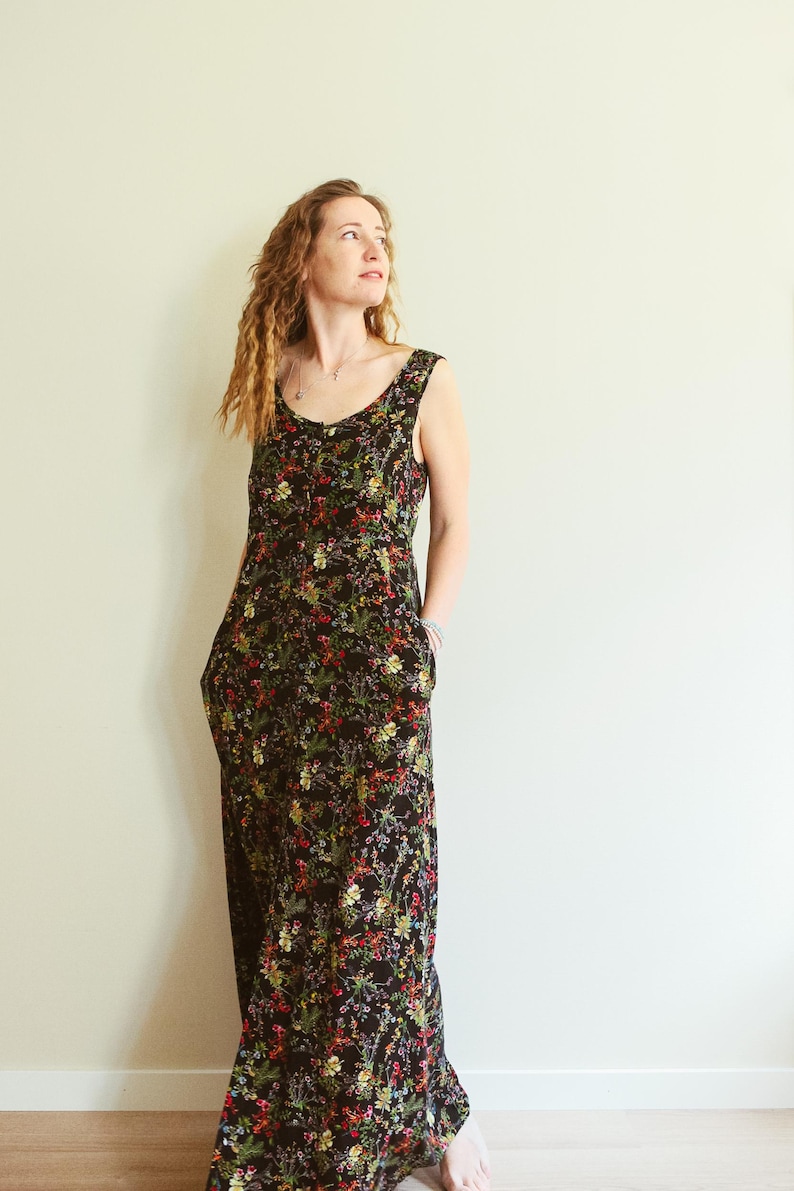 May include: A sleeveless, floor-length dress with a black background and a vibrant floral pattern in red, yellow, and green. The dress has a scoop neckline and pockets. The model is wearing the dress.