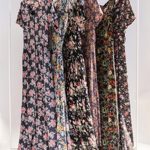 May include: Five floral print dresses hanging on a white clothing rack. The dresses feature various dark backgrounds with colorful floral patterns. Each dress has a different neckline and sleeve style, showcasing a range of designs.