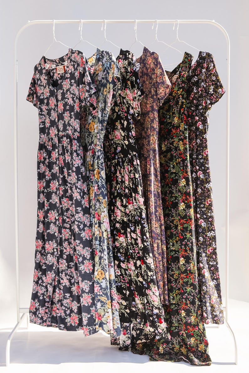 May include: Five floral print dresses hanging on a white clothing rack. The dresses feature various dark backgrounds with colorful floral patterns. Each dress has a different neckline and sleeve style, showcasing a range of designs.