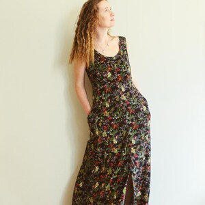 May include: A sleeveless, floor-length dress with a black background and a vibrant floral pattern in red, yellow, and green. The dress features a scoop neckline, pockets, and a front slit. The model is wearing the dress.