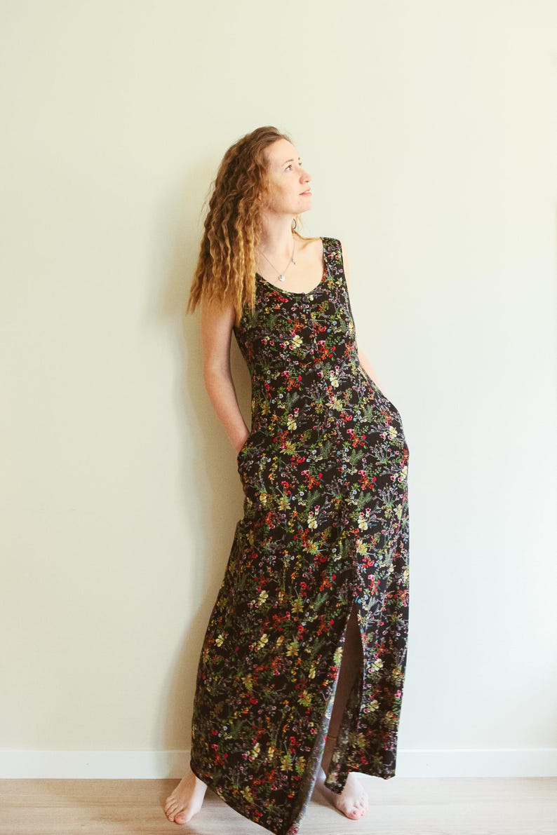 May include: A sleeveless, floor-length dress with a black background and a vibrant floral pattern in red, yellow, and green. The dress features a scoop neckline, pockets, and a front slit. The model is wearing the dress.