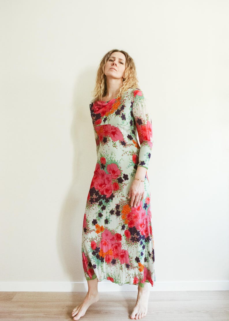 May include: A long-sleeved maxi dress with a floral print in shades of pink, orange, green, and black. The dress has a cowl neck and a fitted silhouette.