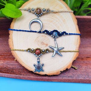 Op de afbeelding: Drie handgemaakte vriendschapsarmbanden met verschillende bedels. De bovenste armband is beige met bruine kralen en een halve maan bedel. De middelste armband is blauw met een blauwe steen en een zeester bedel. De onderste armband is groen met rode kralen en een schildpad bedel.