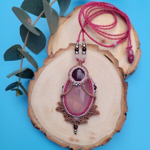 Può includere: Una collana macramè fatta a mano con un grande ciondolo ovale in quarzo rosa. Il ciondolo è incorniciato da un intricato macramè nei toni del marrone e del rosa, impreziosito da piccole perline e una pietra rosso scuro. La collana ha un cordino rosa.