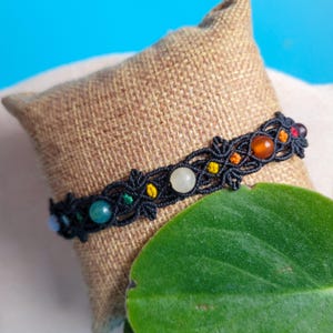 Può includere: Un braccialetto in macramè nero con perline colorate, tra cui verdi, gialle, arancioni e bianche. Il braccialetto è esposto su un cuscino di juta con una foglia verde in primo piano.