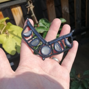 Op de afbeelding: Een zwarte en blauwe macramé ketting met vijf ronde, lichtgrijze stenen. De ketting heeft de vorm van een V en wordt vastgehouden door een hand.